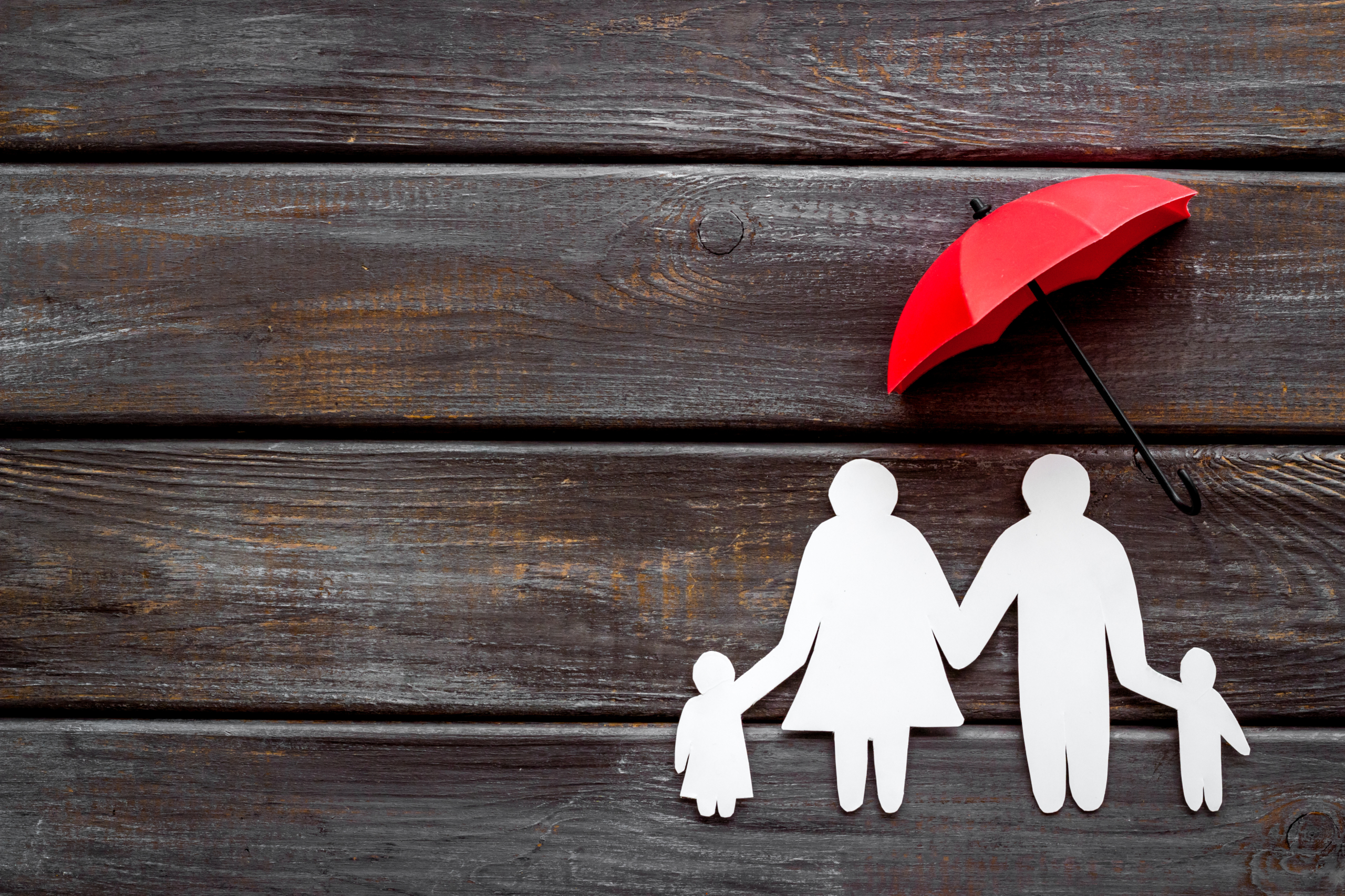 Live insurance concept. Family silhouette protected by umbrella on dark wooden background top-down copy space